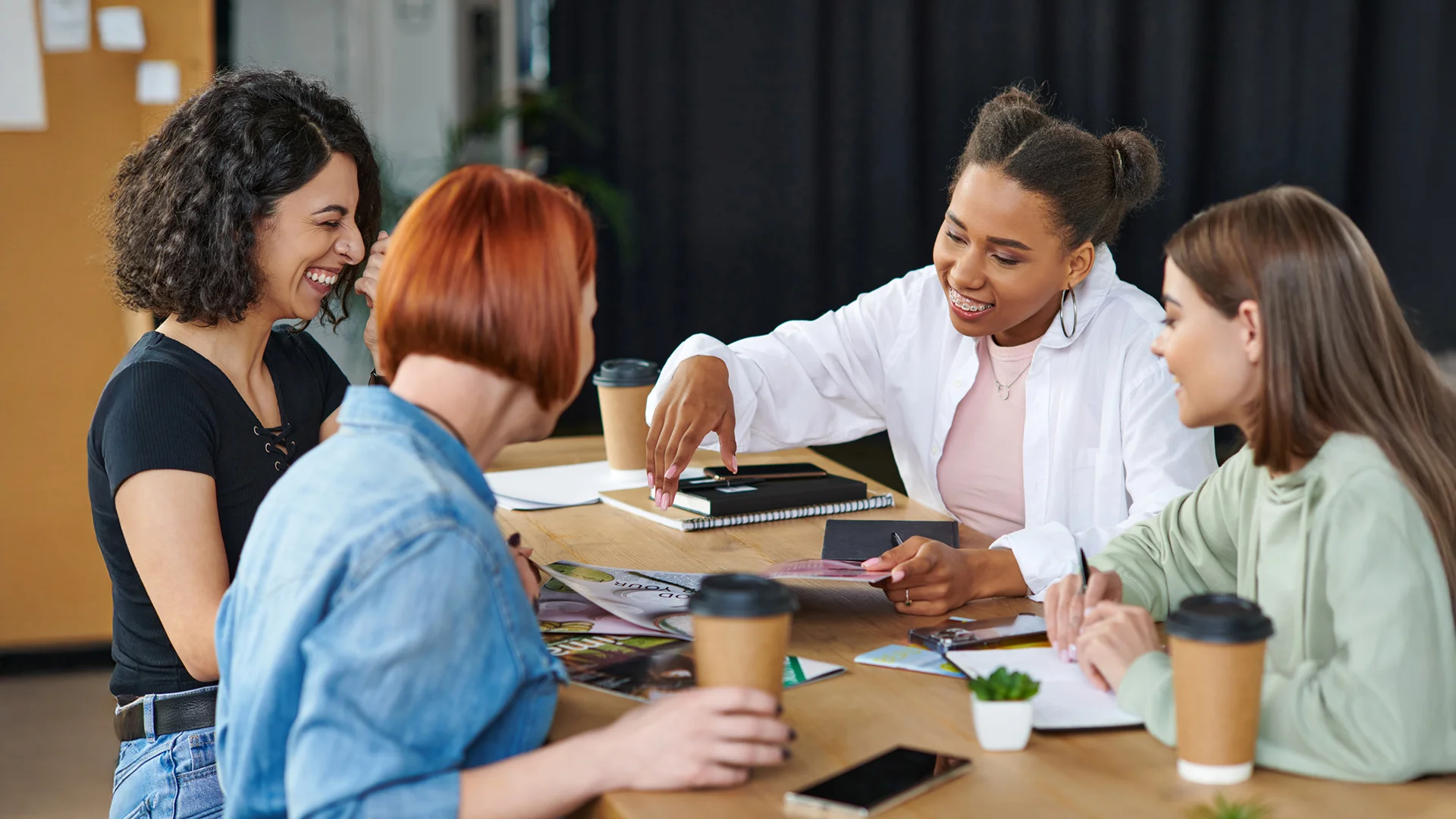 Creative Team Collaborating Over Magazines And Notes In Casual Office Group of diverse young women laughing and sharing ideas around a table with magazines, coffee cups, and notebooks.